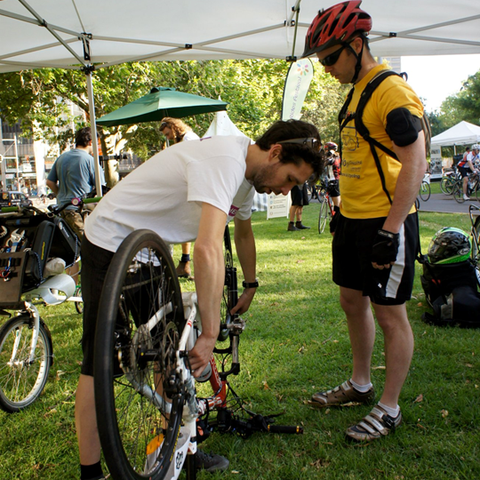 Two men in bike gear and helmets under a shelter with a bike - one is fixing the bike wheel